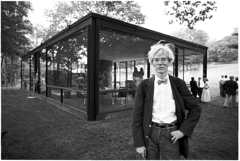 Person with glasses and bow tie standing in front of a modern glass house with trees and people in the background.