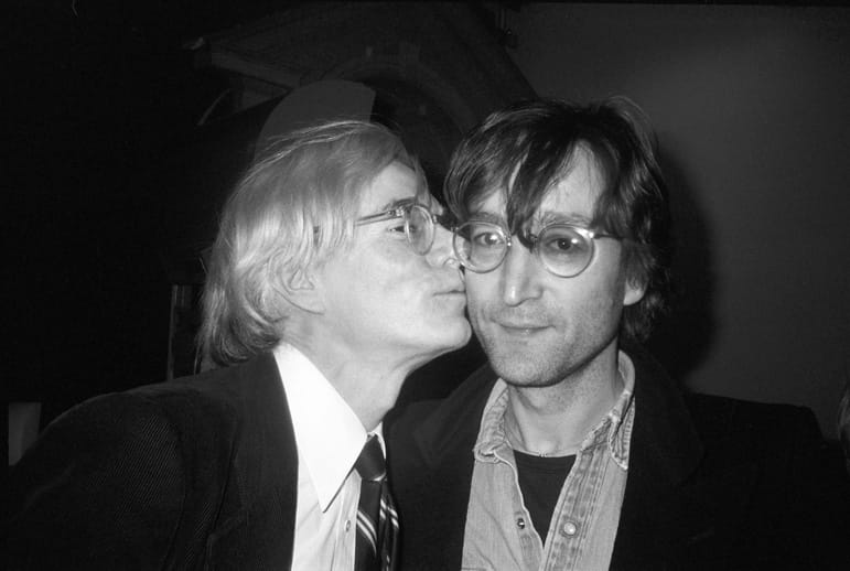 Two men, one kissing the other on the cheek, both wearing glasses and suits, with a dark background.
