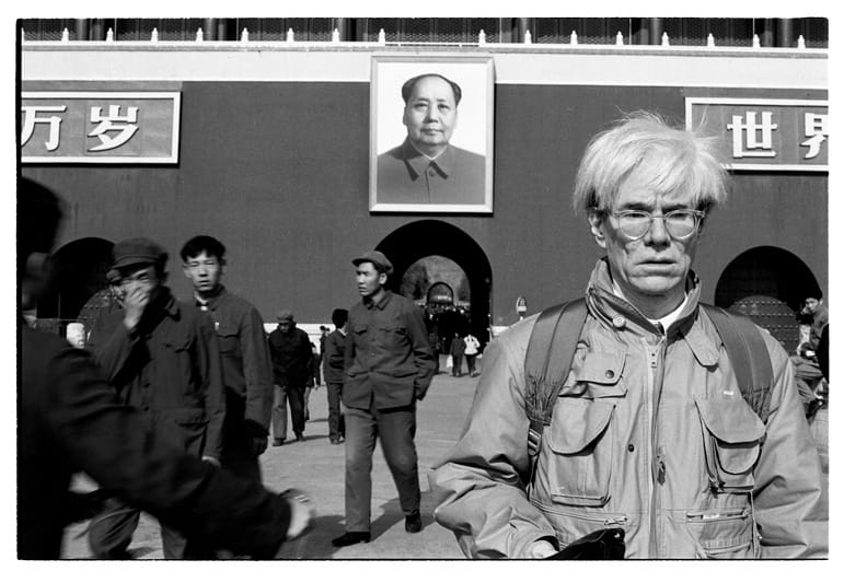 Person standing in front of a building with a giant portrait of Mao Zedong, surrounded by people walking.