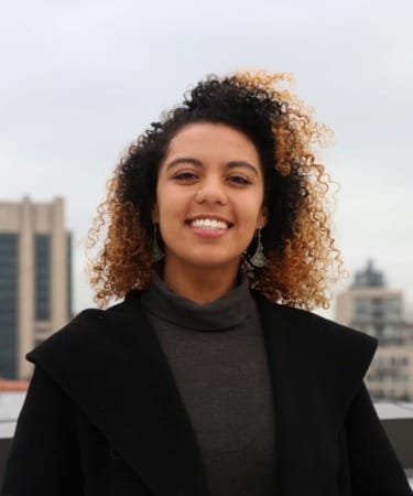 Smiling person with curly hair wearing a dark coat and turtleneck on an urban rooftop with buildings in the background.