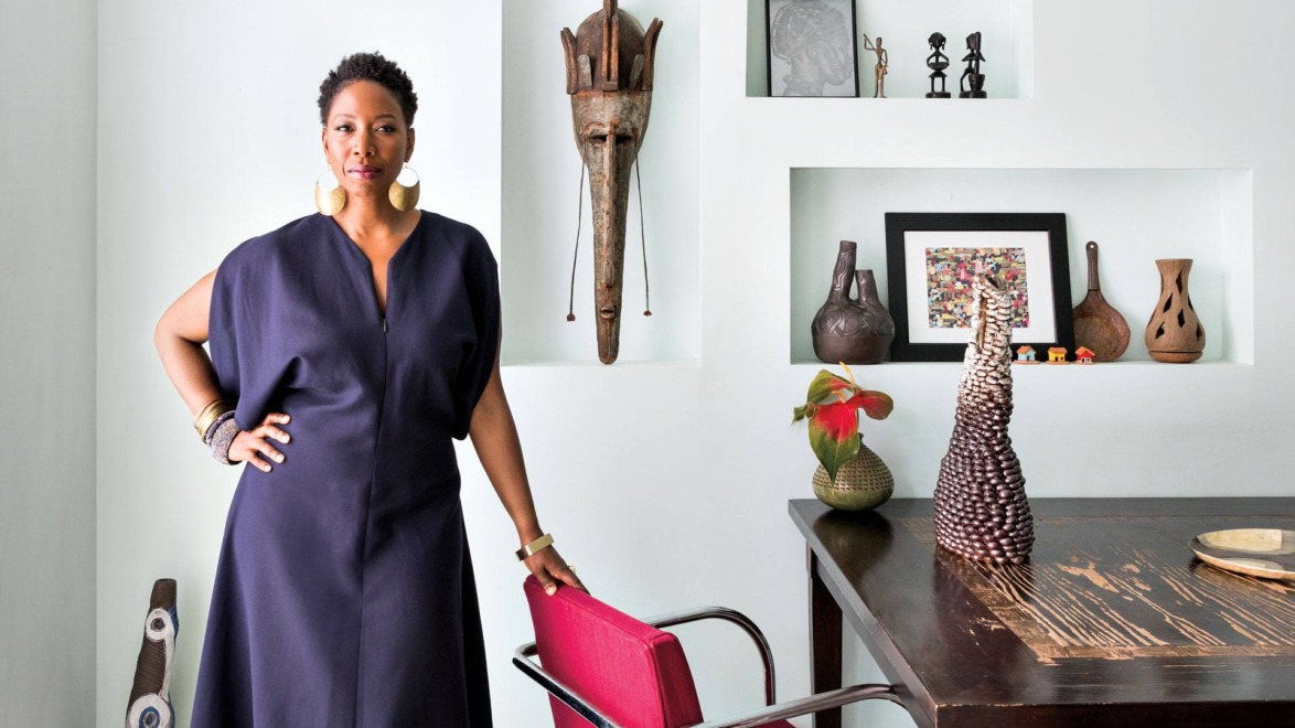 Person in a blue dress stands by a table in a room with cultural artifacts and art on display.