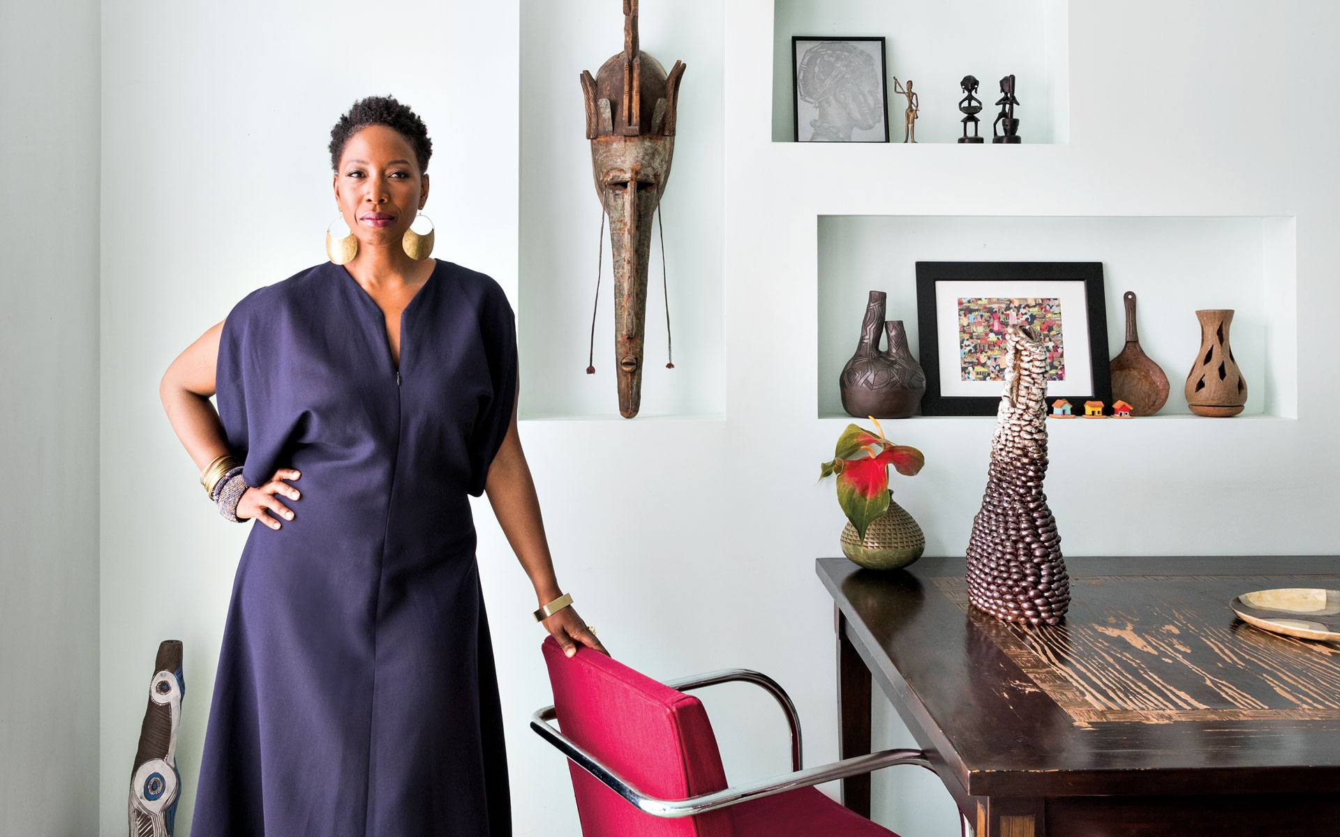Person in a blue dress stands by a table in a room with cultural artifacts and art on display.