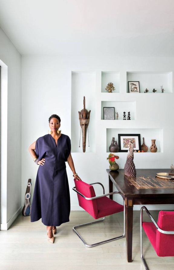 Woman standing in a modern dining room with wall art, decorative shelves, and a wooden table with red chairs.