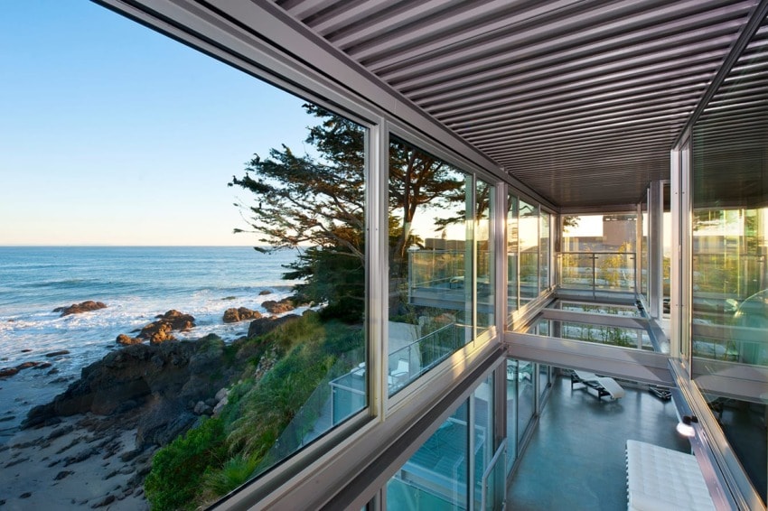 Modern oceanfront house with large glass windows overlooking rocky shore and trees at sunset.