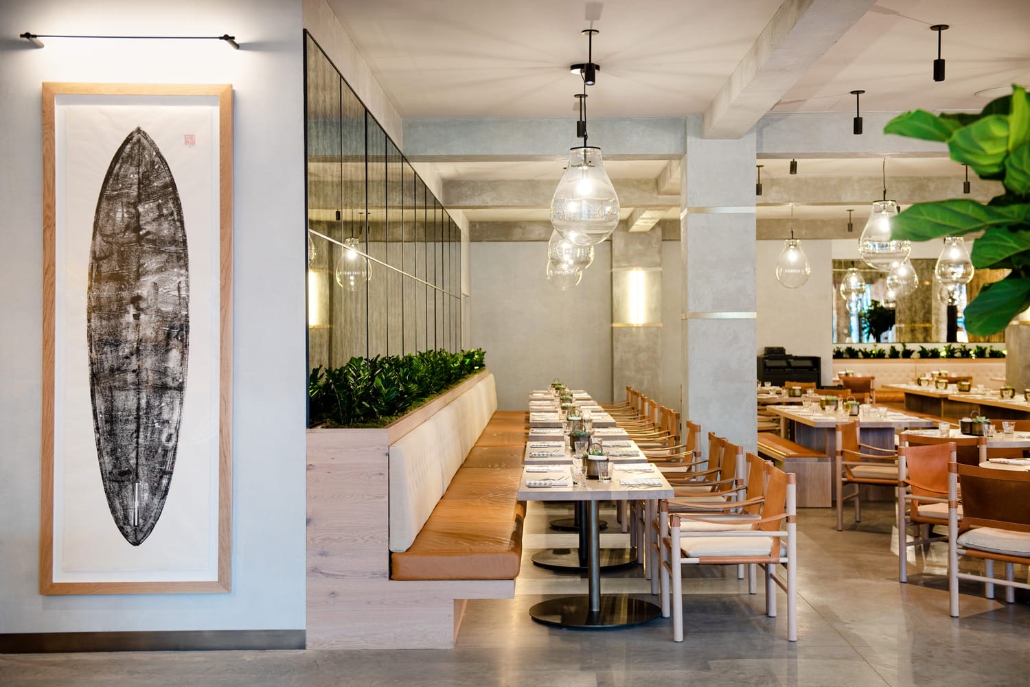 Modern restaurant interior with wooden tables, cushioned seating, hanging lights, and a surfboard artwork displayed on the wall.