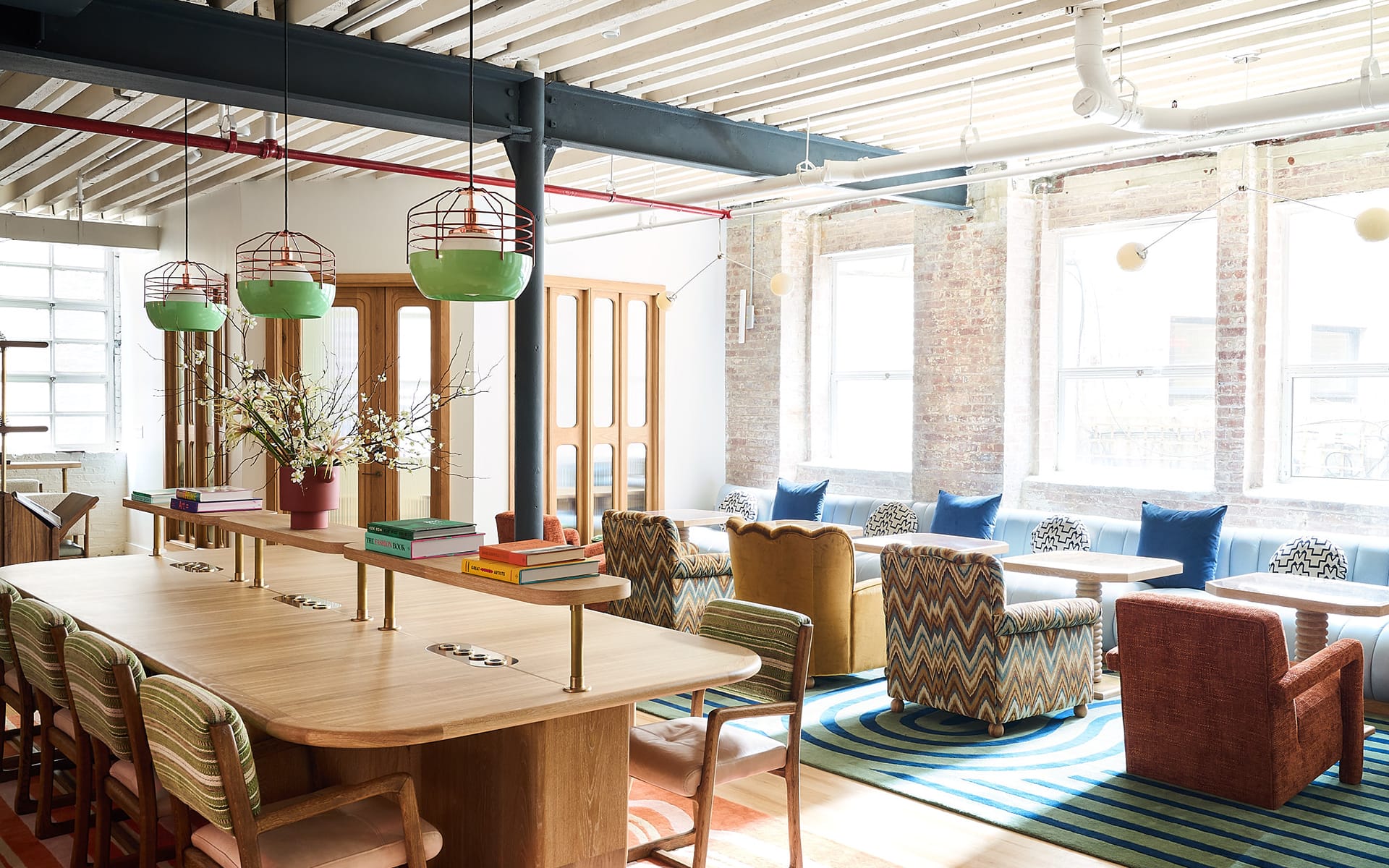 Bright modern coworking space with wooden tables, colorful chairs, large windows, and hanging green lights.