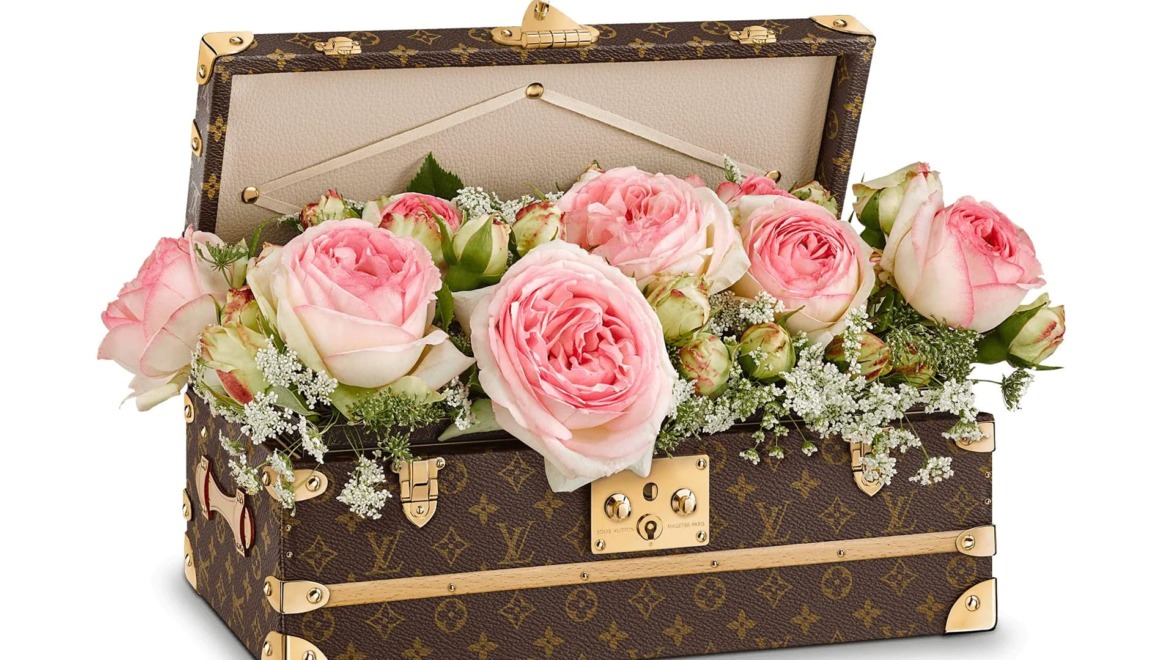 Open decorative brown and gold box filled with pink roses and delicate greenery, arranged beautifully.