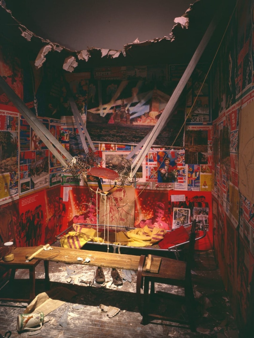 Room with torn posters, hanging strings, and debris, featuring a wooden bench and red walls with peeling paint.