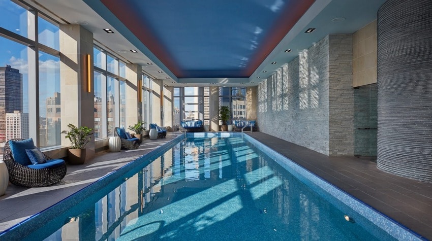 Indoor hotel pool with skylights and city views, surrounded by chairs and plants, featuring stone and wood accents.