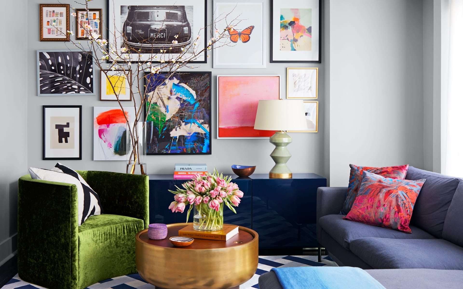 Colorful living room with an art gallery wall, green velvet chair, blue sofa, gold coffee table, and pink tulips in a vase.