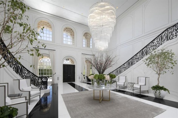 Luxury foyer with grand chandelier, dual staircases, elegant decor, large windows, and potted plants.