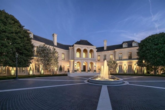 Large elegant mansion with a grand entrance and a fountain in the foreground, surrounded by trees at dusk.
