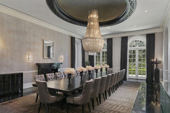Elegant dining room with long table, tufted chairs, large chandelier, and arched windows overlooking a lush garden.
