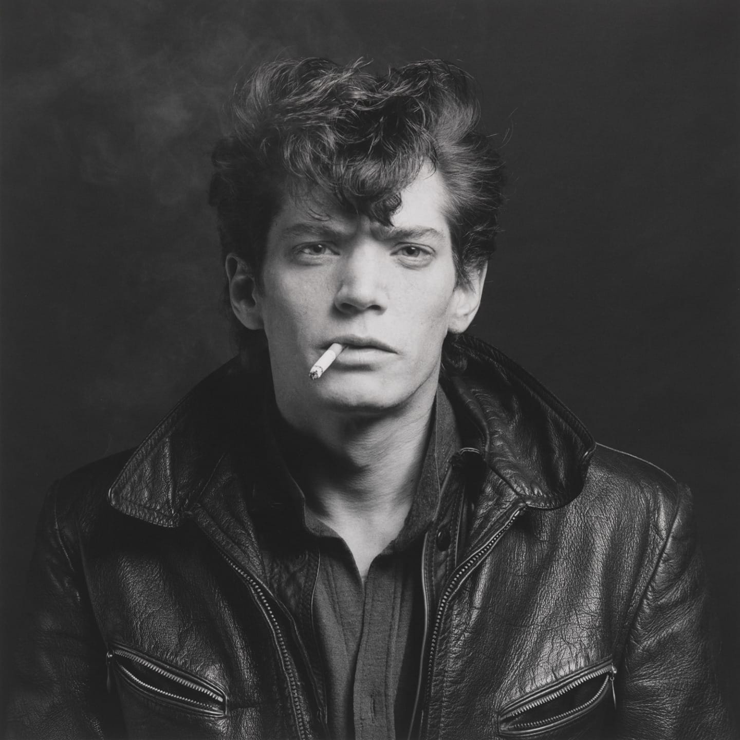 Person with a cigarette wearing a leather jacket, gazing forward with a neutral expression. Black and white photograph.