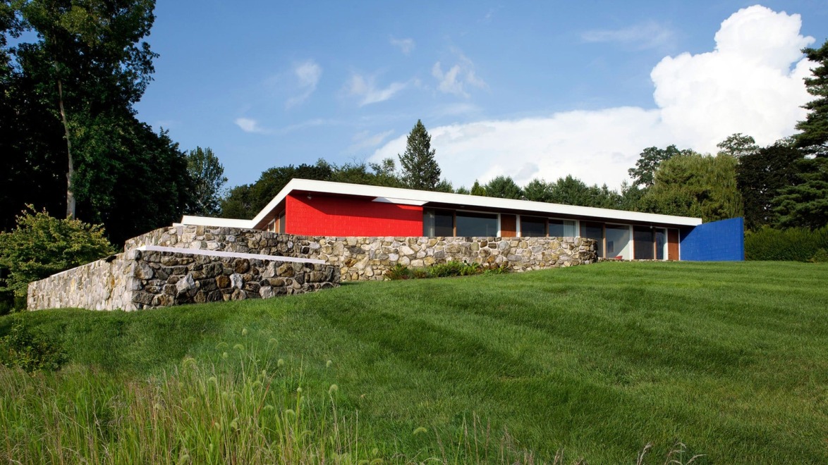 Modern house with vibrant red and blue accents, surrounded by a lush green lawn and stone wall in a serene natural setting.