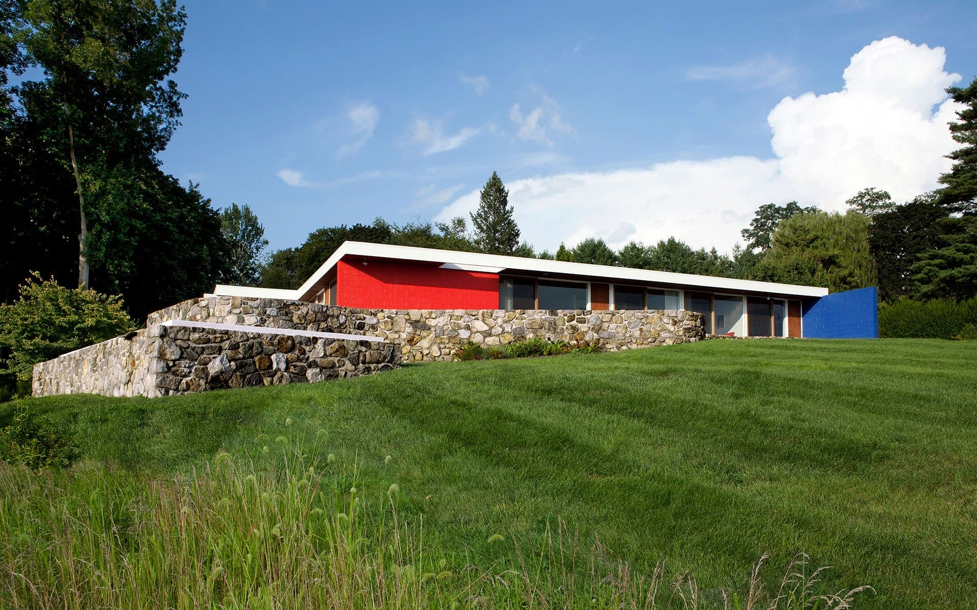 Modern house with vibrant red and blue accents, surrounded by a lush green lawn and stone wall in a serene natural setting.