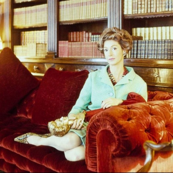 Woman in a green dress sitting on a red velvet couch in a room with wooden bookshelves filled with leather-bound books.