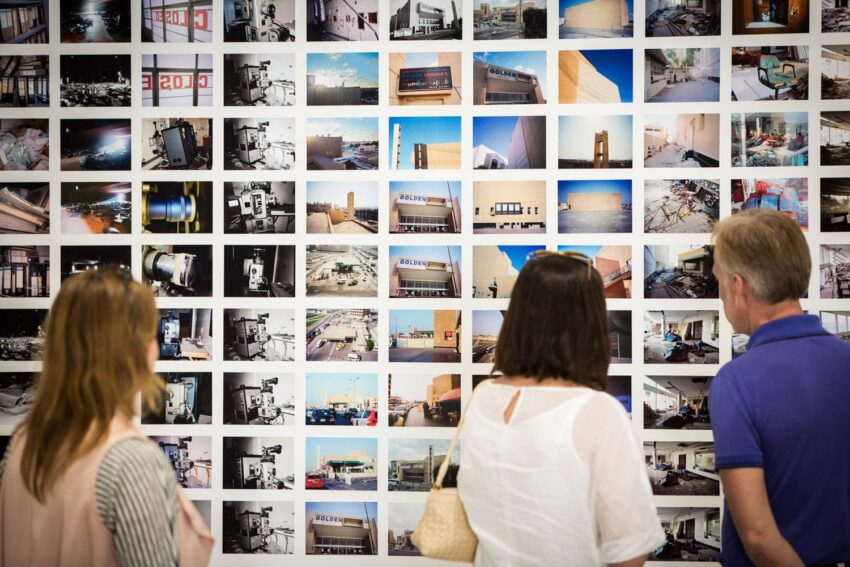 People viewing a wall of various photographs displayed in a gallery exhibition setting.