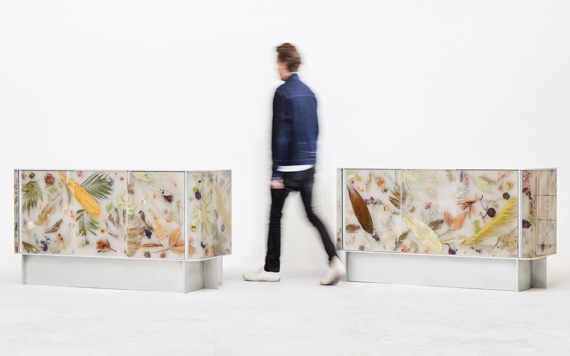 Person walking past two decorative sideboards with assorted natural elements encased in resin, against a plain background.