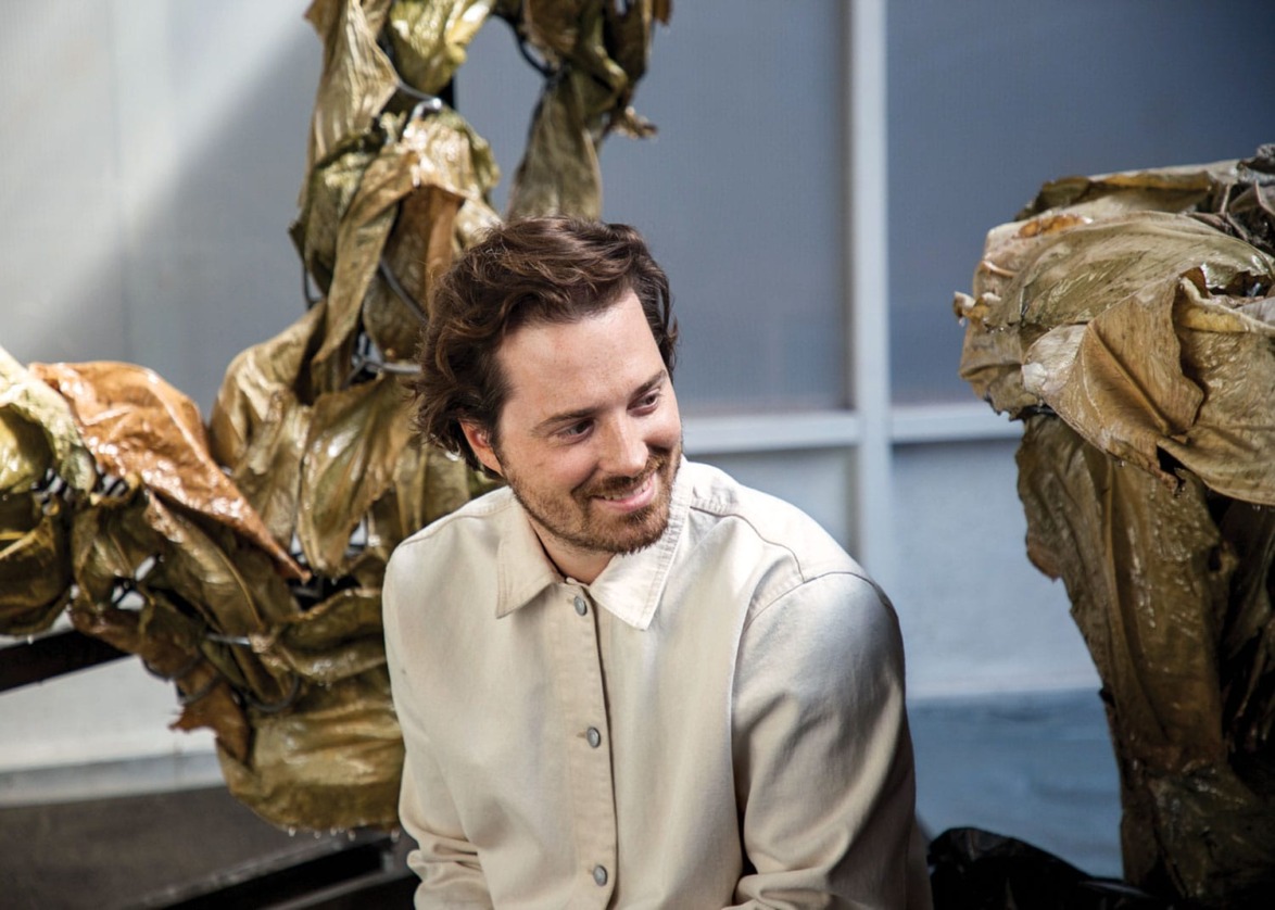 Man sitting and smiling, wearing a beige jacket, surrounded by large abstract sculptures indoors.