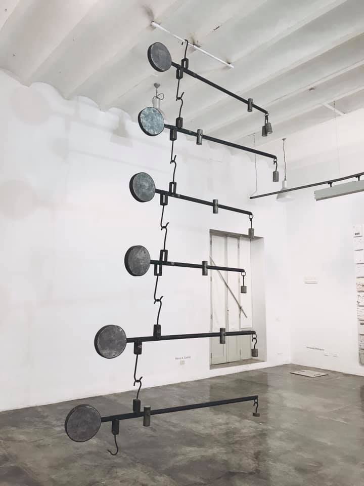 Abstract metal sculpture with circular plates suspended from a white ceiling in a minimalistic gallery setting.