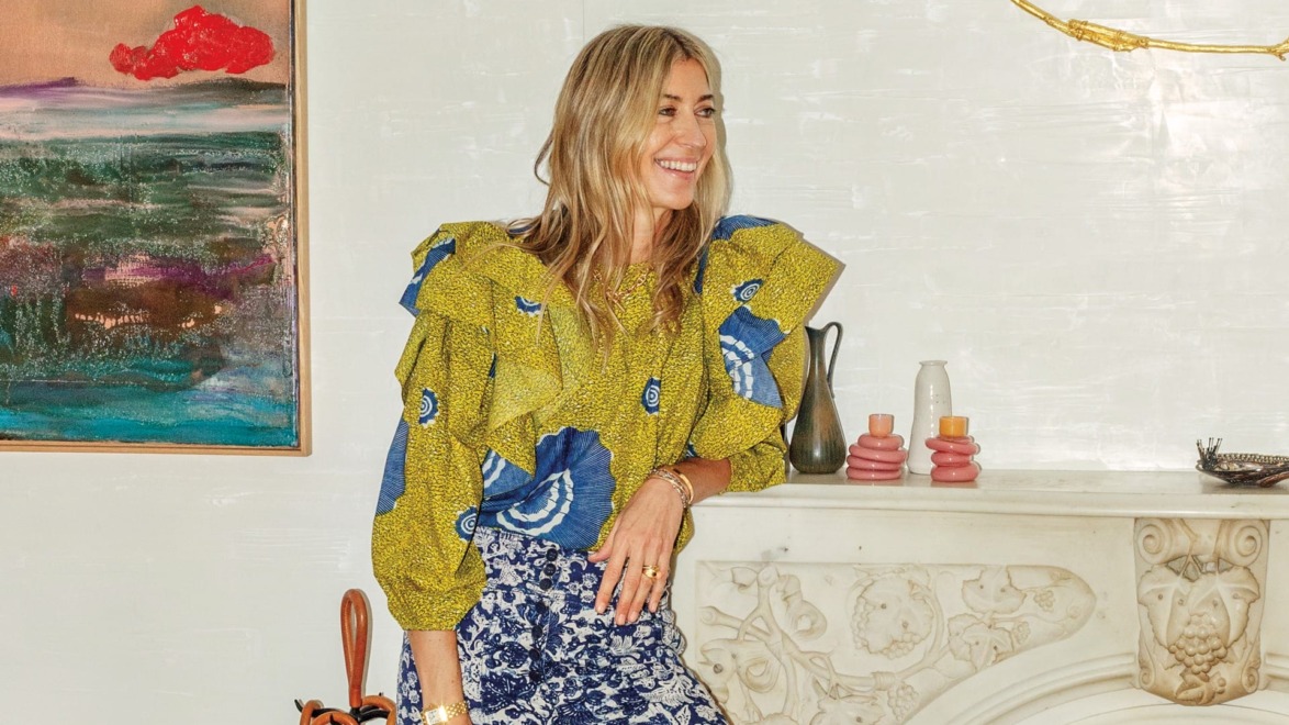 Person with long blonde hair, wearing a patterned blouse and skirt, standing by a fireplace, colorful painting in the background.