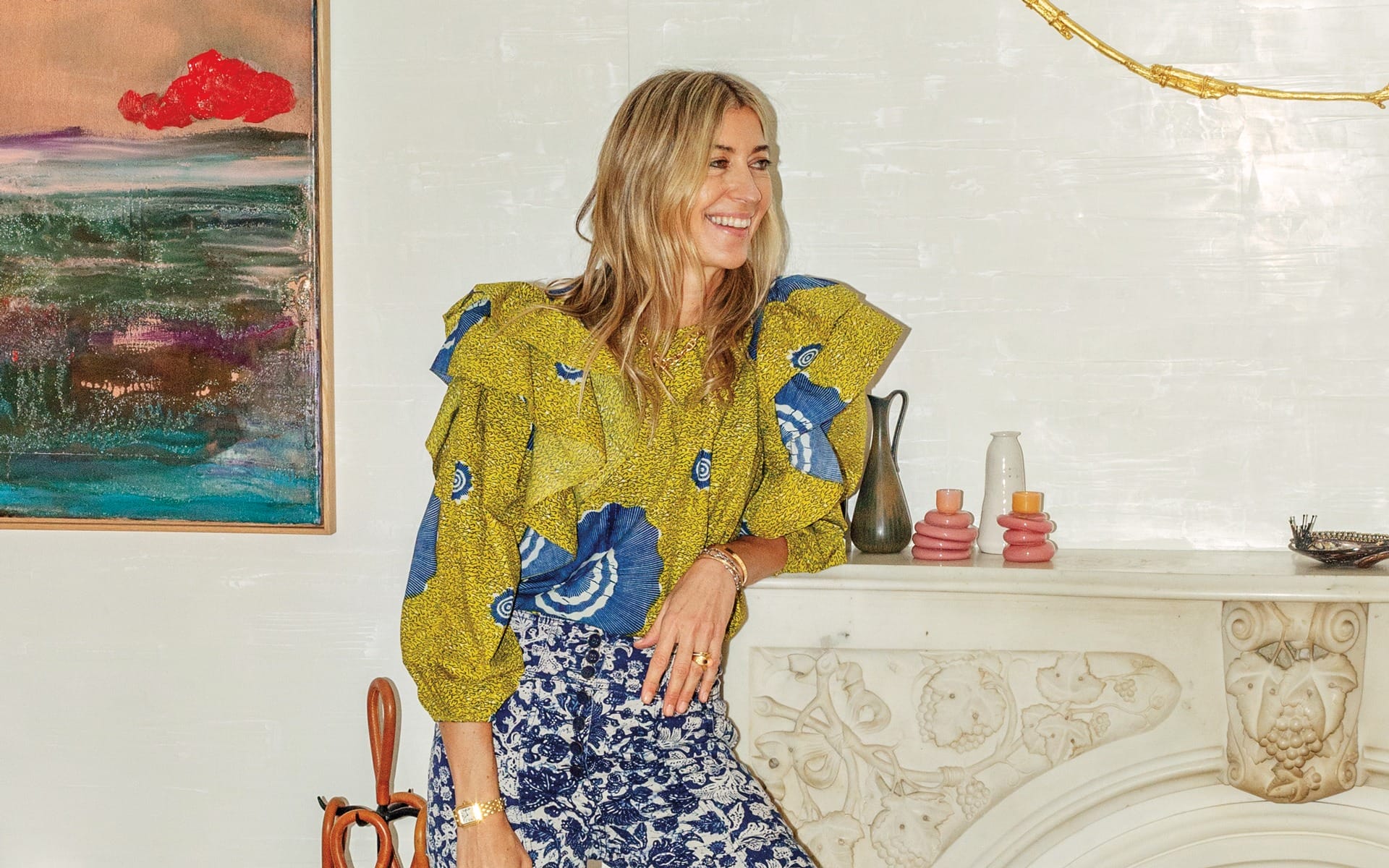 Person with long blonde hair, wearing a patterned blouse and skirt, standing by a fireplace, colorful painting in the background.