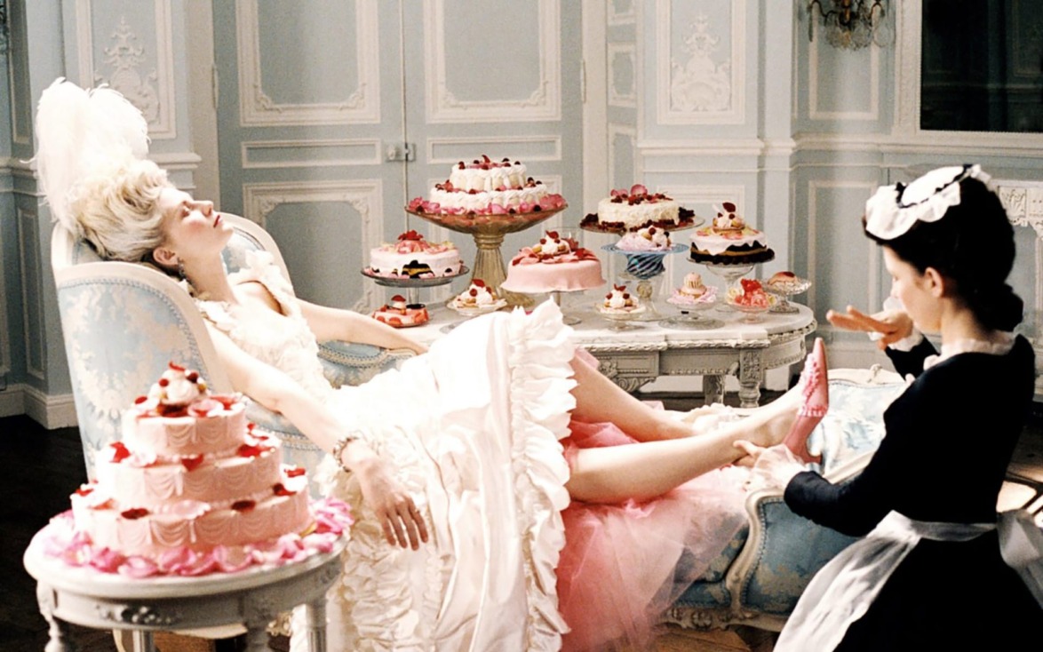Woman in an ornate dress relaxing on a chaise surrounded by lavish cakes, with an attendant adjusting her shoe nearby.