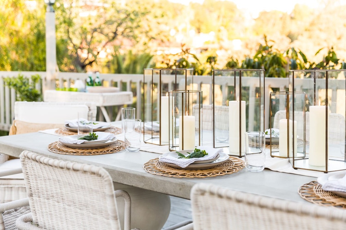 Outdoor dining table set with wicker placemats, white plates, glass lanterns, and greenery, surrounded by chairs and trees.