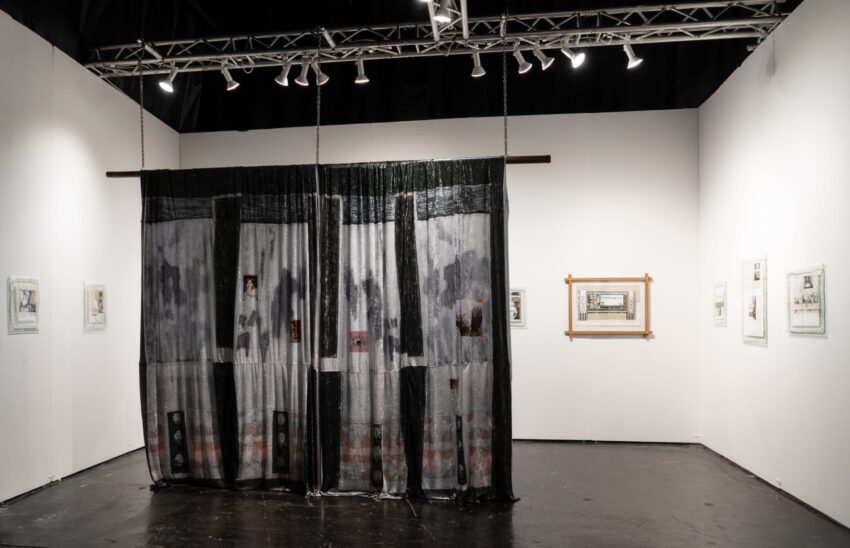 Art installation with a large hanging textile in a gallery, surrounded by framed artworks on white walls and spotlighting above.