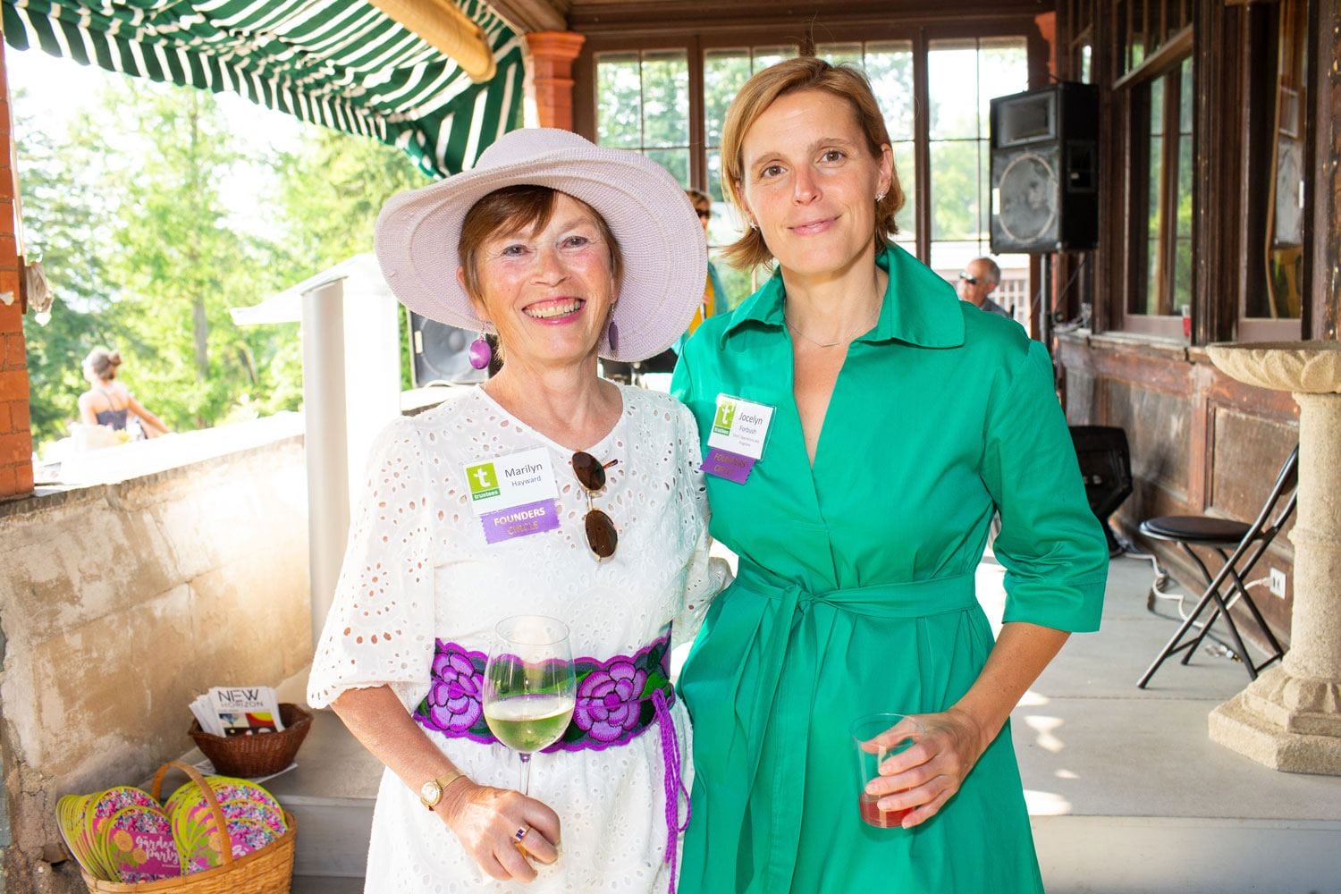 Two women smiling at an outdoor event, one in a white dress and hat, the other in a green dress, holding drinks.