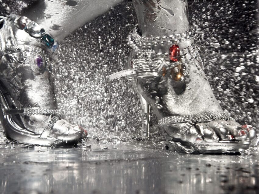 High-heeled sandals adorned with colorful gems on feet covered in silver body paint, amid a splash of shimmering liquid.