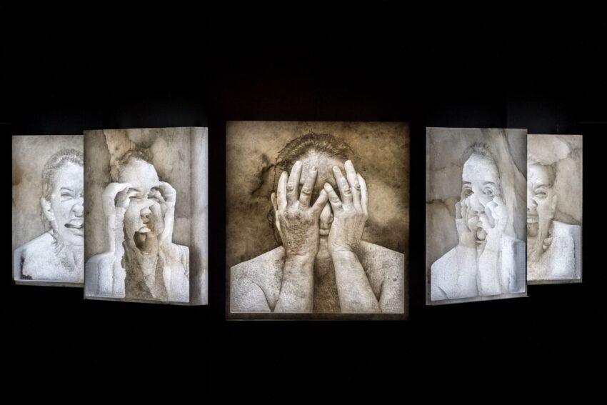 Artistic installation of expressive portraits depicting a person covering their face and peeking through fingers against a dark background.