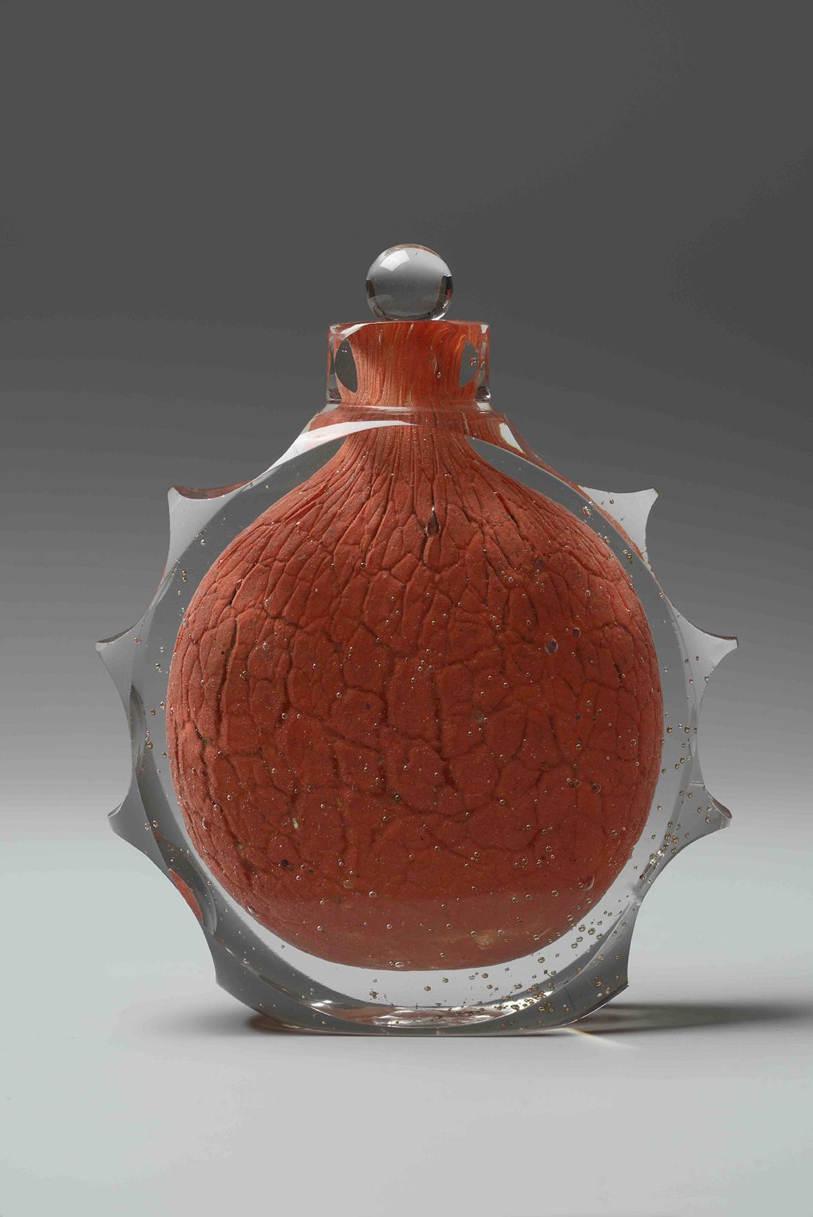 Decorative glass bottle with a round, textured red center and spiked edges against a gray background.