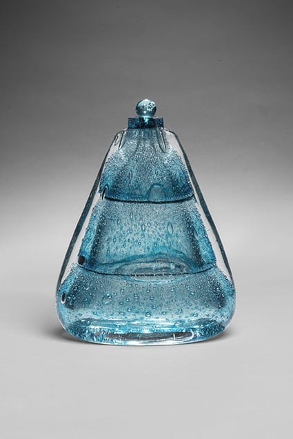 Blue glass sculpture with a rounded top, featuring air bubbles, against a gradient gray background.