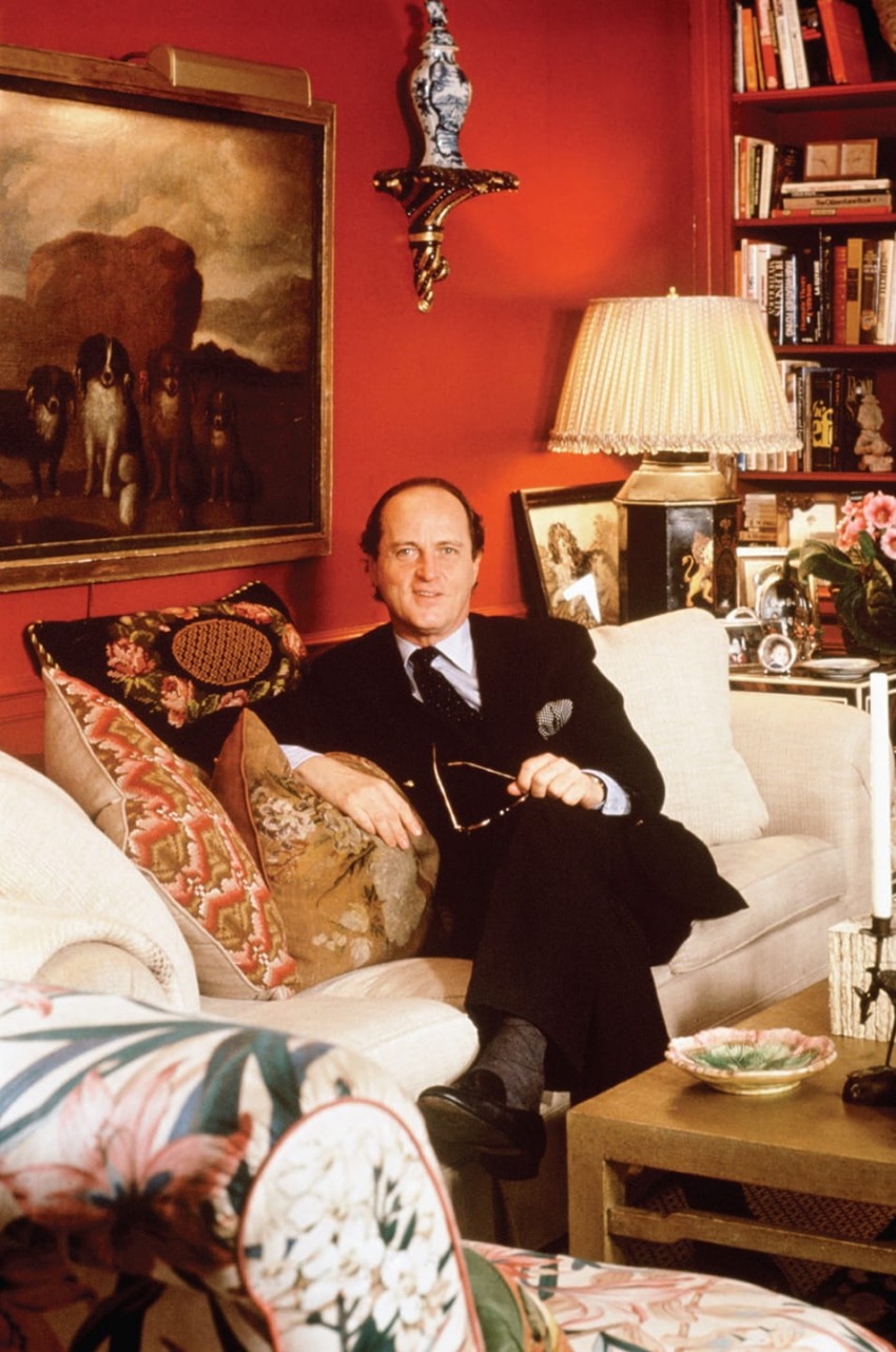 Man sitting in a stylish, art-filled living room with colorful cushions and lamp, surrounded by books and a dog painting.