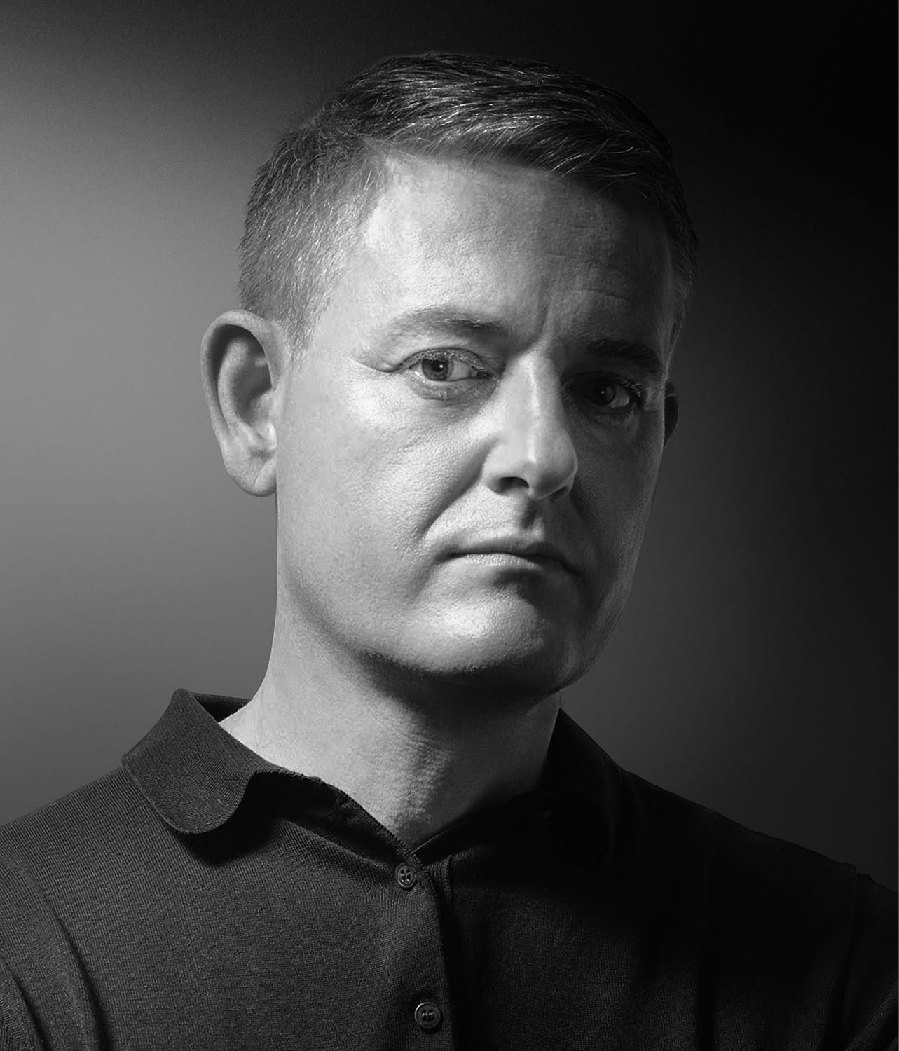 Black and white portrait of a person with short hair, wearing a polo shirt, looking at the camera against a gradient background.
