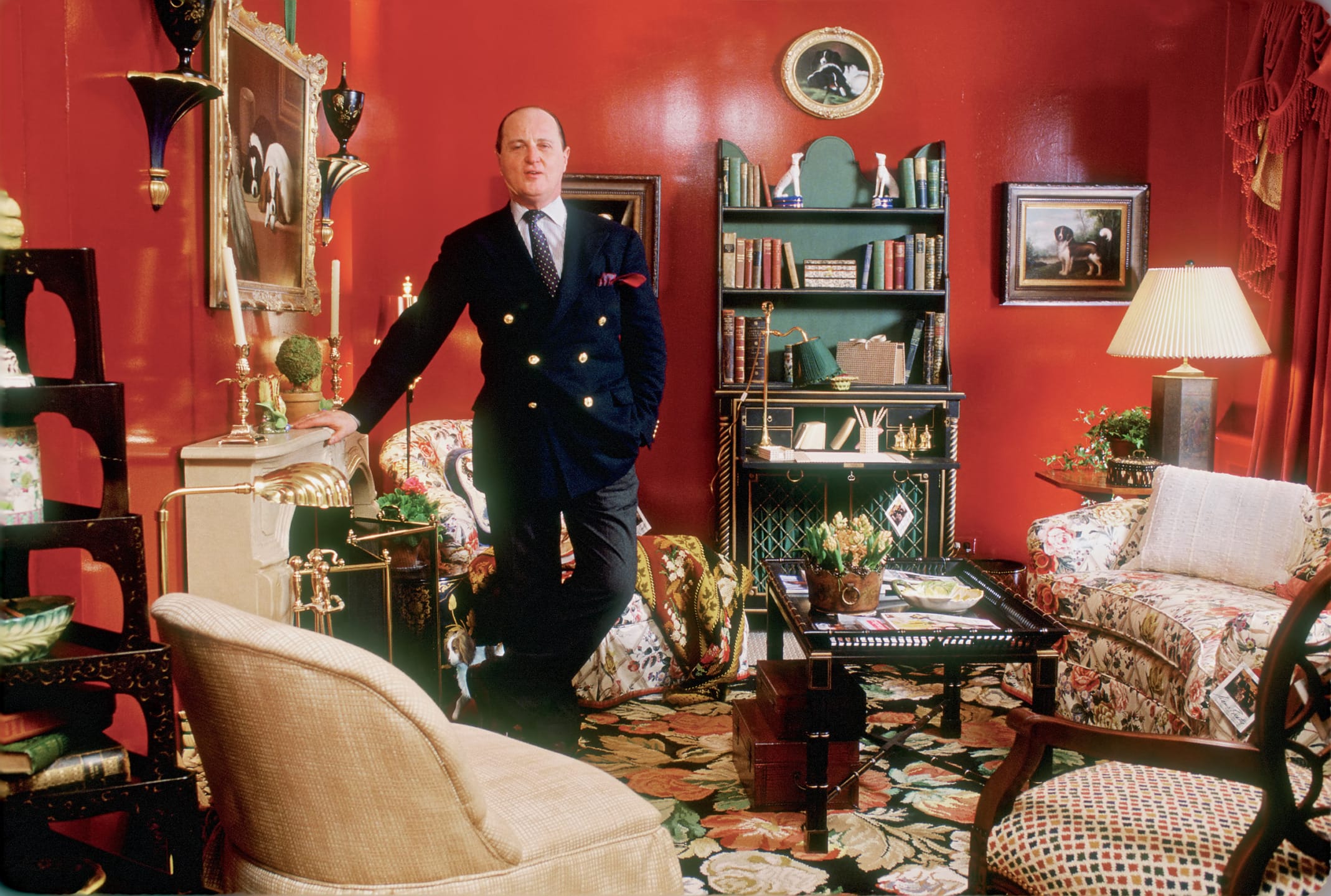 Man in stylish living room with red walls, patterned carpet, and vintage decor elements, standing next to a bookshelf.