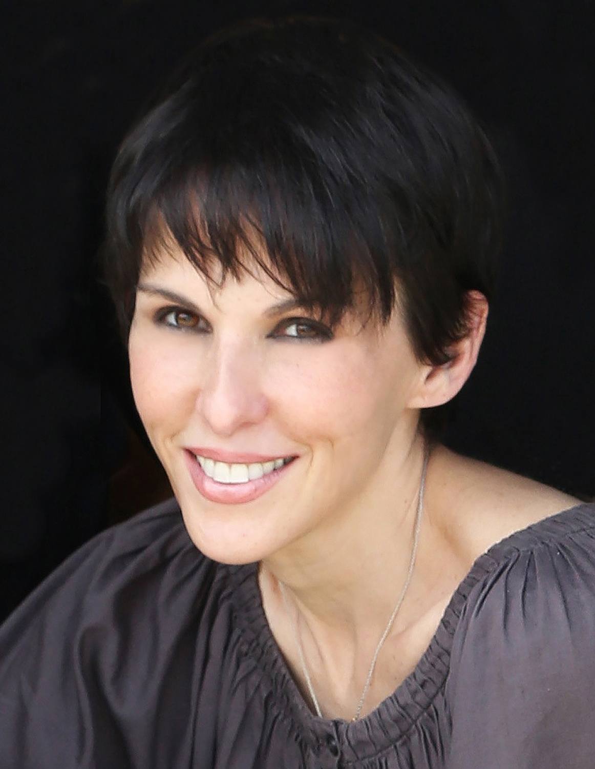 Person smiling with short dark hair, wearing a gray blouse, against a black background.