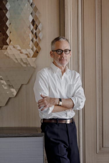 Man with glasses and white shirt leaning against a wall, standing next to a modern decorative piece in a wooden room.