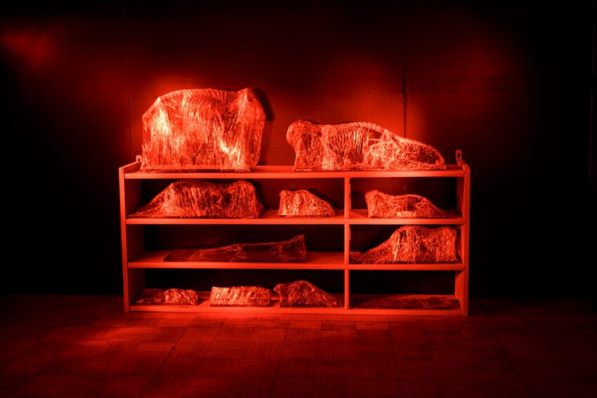 Shelf with abstract sculptures wrapped in reflective material, illuminated by red lighting in a dark room.