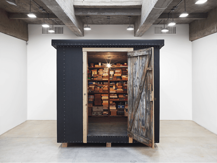 Small wooden storage room with an open door displaying shelves filled with various boxes under bright ceiling lights.