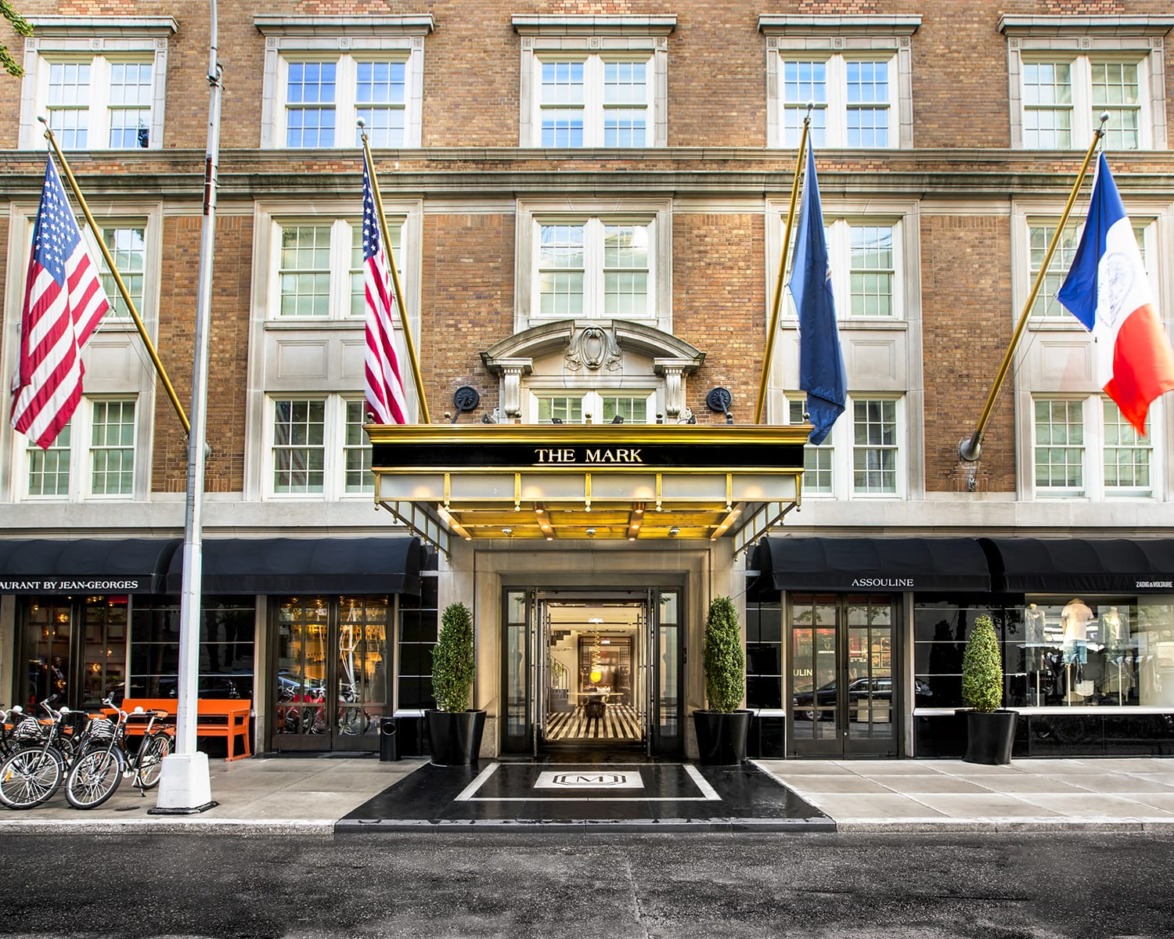 Exterior view of a luxury hotel entrance with flags, showing a classic brick facade and elegant awning over the doorway.
