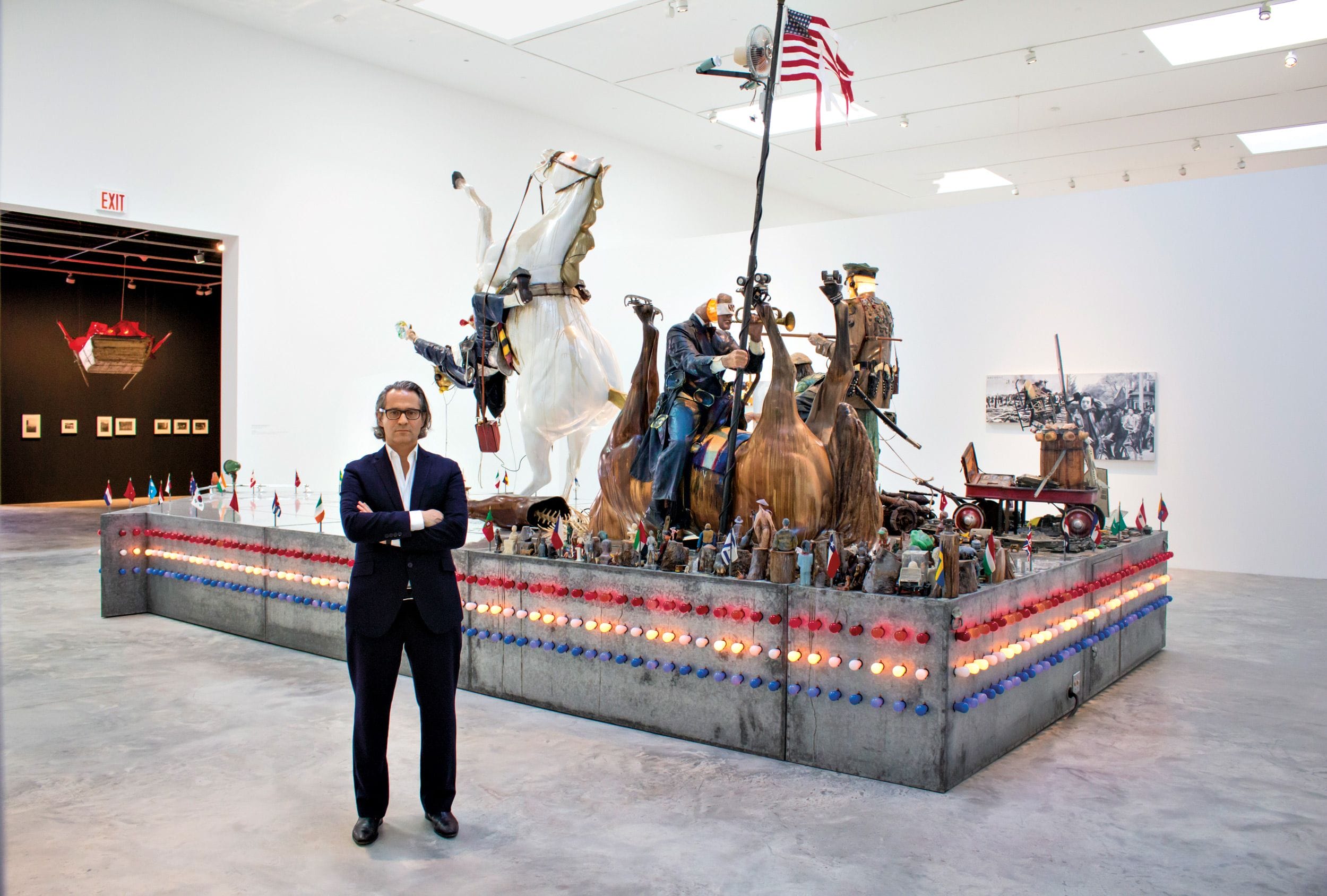 Man standing in front of a large, patriotic art sculpture with historical figures and flags in a modern gallery setting.