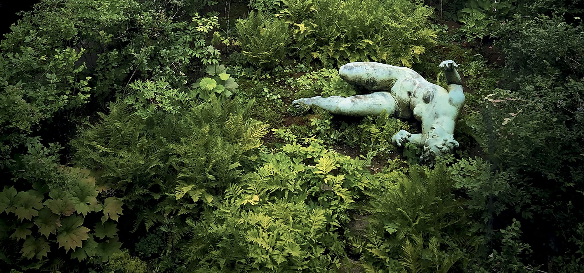 Statue of a reclining figure partially covered by lush greenery in a garden setting.