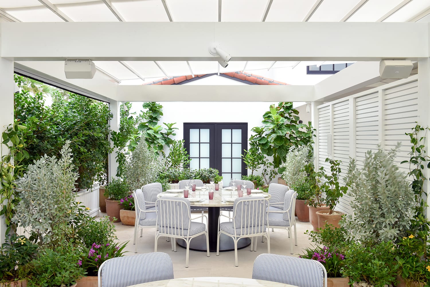 Outdoor patio with dining table, chairs, and lush greenery under a pergola with a white roof and modern design elements.