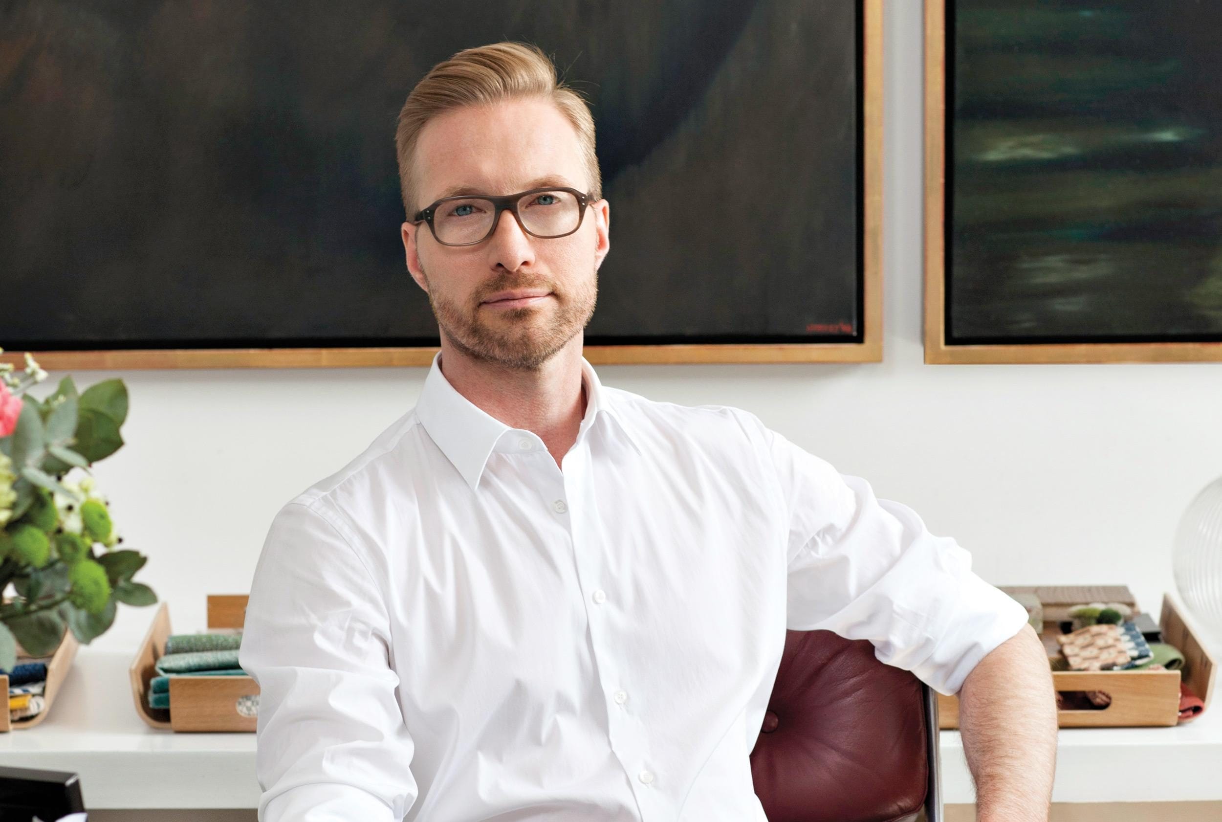 Man in a white shirt and glasses sitting in front of paintings, with a confident expression.