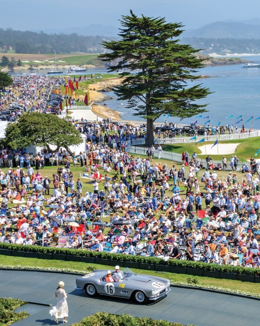 Classic car driving at a crowded outdoor automotive event near the ocean with people enjoying the scenery and festivities