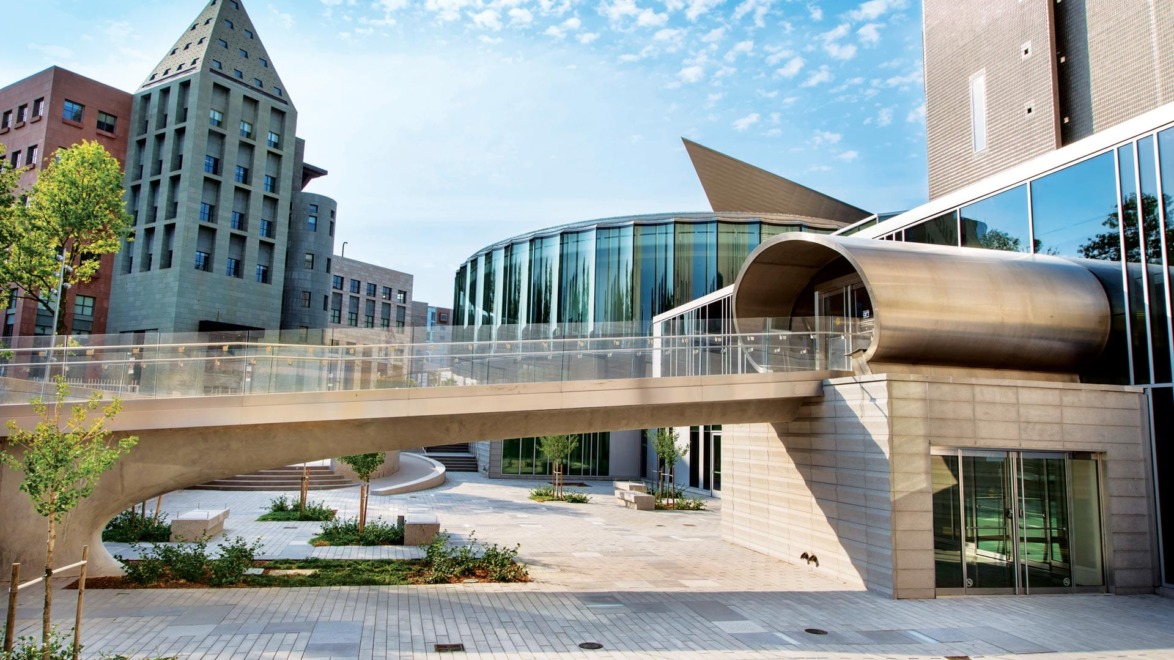 Modern architectural building with large glass windows, sleek bridge, and unique geometric shapes under a clear blue sky.