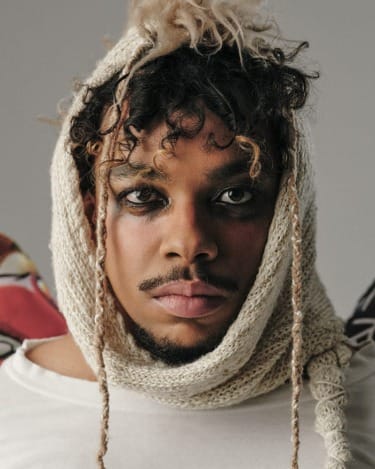 Person wearing a knitted hooded scarf, with a neutral expression and curly hair, looking directly at the camera.